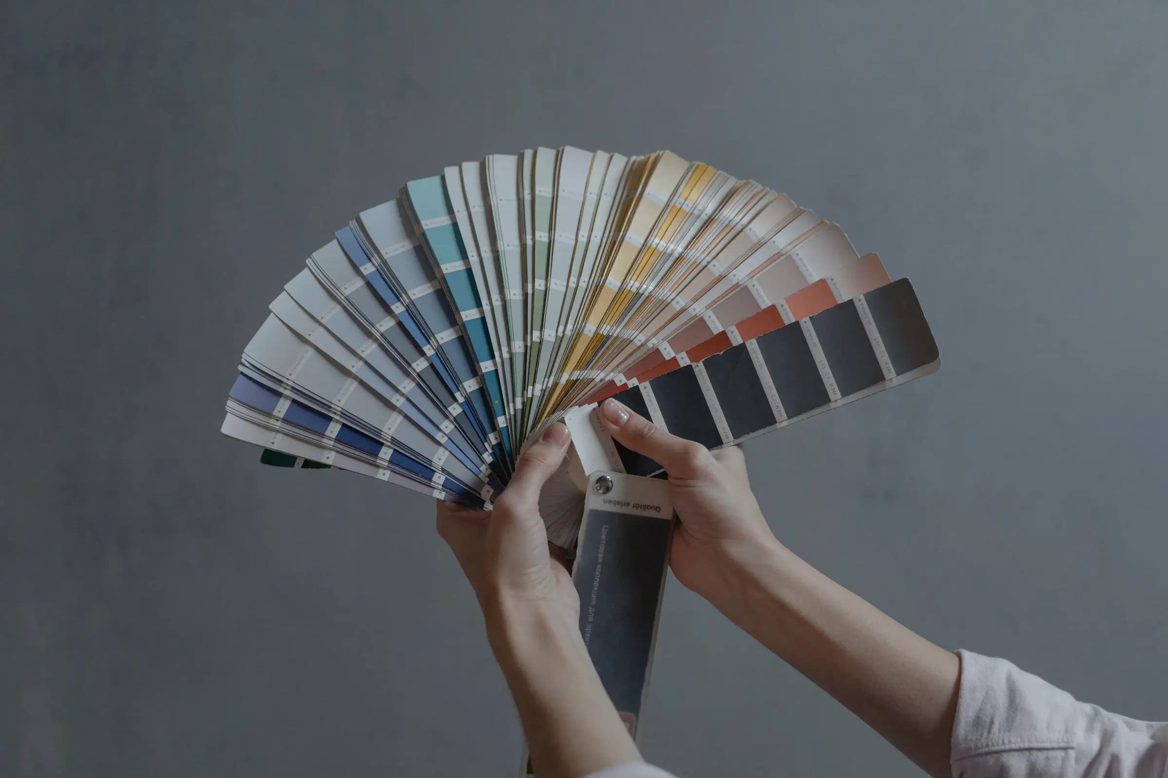 Manos que abren un abanico de muestras de color frente a un muro gris.